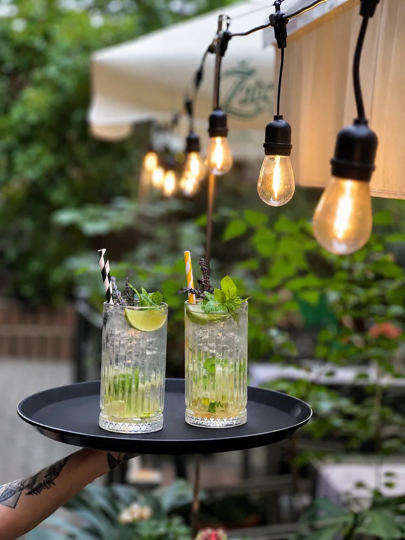 Two drinks with lime and mint on a tray beneath courtyard string lights