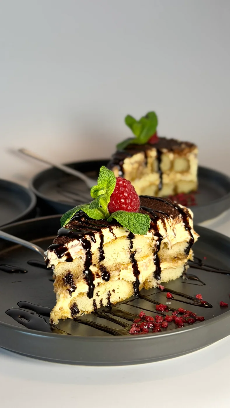 Two portions of tiramisu with chocolate sauce, raspberries and fresh mint on dark plates